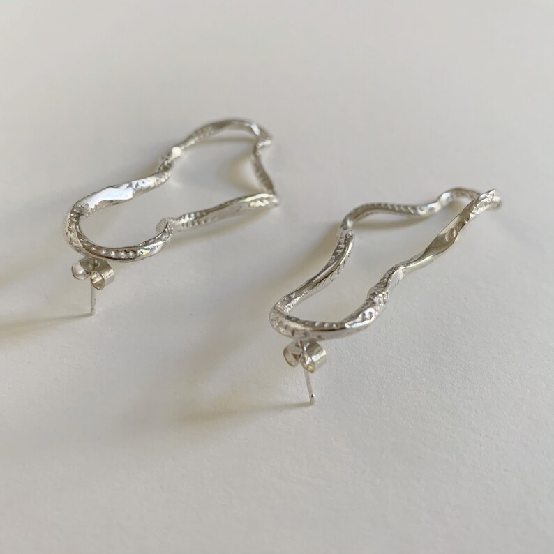 May include: A pair of silver earrings with a unique, wavy, organic shape. The earrings are made of a textured metal and have a post back.