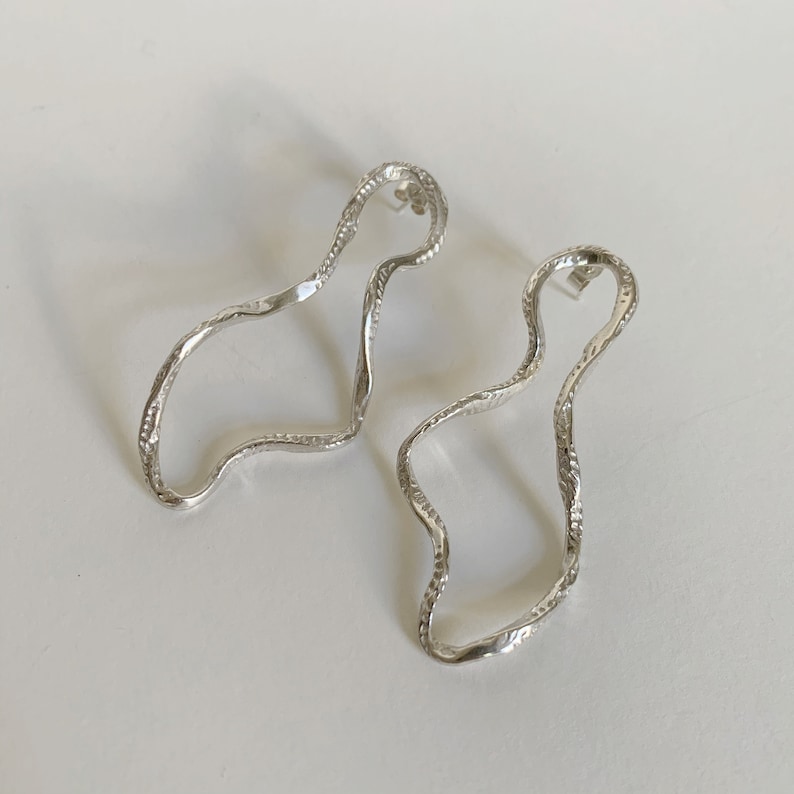 May include: A pair of silver earrings with a wavy, organic shape. The earrings are made of a textured metal and have a simple, modern design.