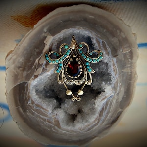 May include: Vintage gold-toned brooch with a central oval garnet stone, surrounded by turquoise and pearl accents. The brooch has an ornate, symmetrical design, and is set against a geode background.
