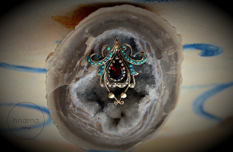 May include: Vintage gold-toned brooch with a central oval garnet stone, surrounded by turquoise and pearl accents. The brooch has an ornate, symmetrical design, and is set against a geode background.