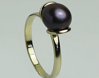 Puede incluir: Un anillo de oro con una gran perla de color púrpura oscuro. La banda del anillo es de oro pulido, curvándose para sostener la perla. La perla es redonda y lisa, con un brillo sutil. Una joya clásica.