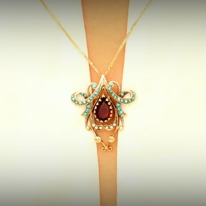 May include: A gold-toned pendant necklace featuring a teardrop-shaped, deep red gemstone at its center. The pendant is adorned with turquoise-colored stones and intricate gold filigree detailing. The necklace is suspended from a delicate gold chain.