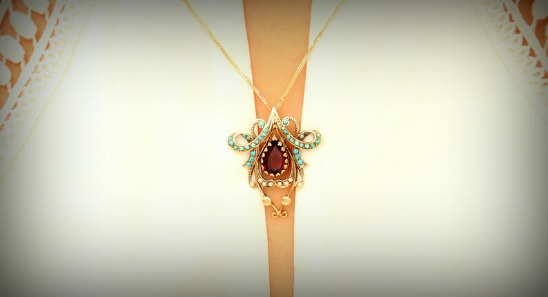 May include: A gold-toned pendant necklace featuring a teardrop-shaped, deep red gemstone at its center. The pendant is adorned with turquoise-colored stones and intricate gold filigree detailing. The necklace is suspended from a delicate gold chain.