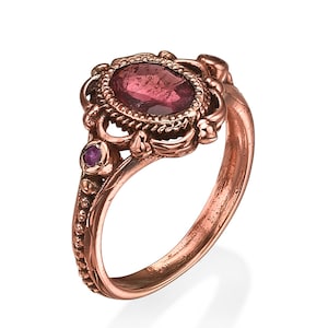 May include: A rose gold ring featuring an oval, reddish-pink gemstone at the center, surrounded by an ornate, textured setting. A smaller, round, reddish-purple gemstone is set on the side. The ring has a detailed, vintage-inspired design.