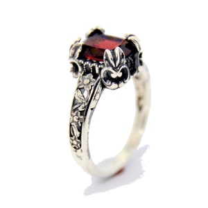 May include: A silver ring with a rectangular, deep red gemstone. The ring features intricate floral and leaf detailing on the band and around the gemstone setting. The design has a vintage or antique aesthetic.