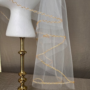 May include: White bridal veil with a gold beaded trim. The veil is displayed on a gold stand.