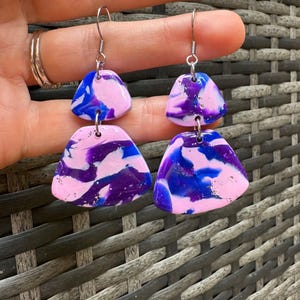 Puede incluir: Un par de pendientes colgantes con dos piezas de forma triangular. La pieza superior es rosa con remolinos blancos y azules. La pieza inferior es morada con remolinos blancos y azules. Los pendientes tienen ganchos plateados.