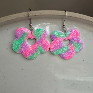 Puede incluir: Un par de pendientes en forma de flor rosa, verde y morado con un corazón recortado. Los pendientes tienen una superficie texturizada y con burbujas.