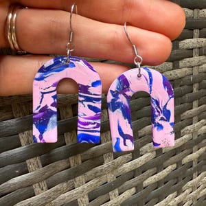 Puede incluir: Un par de pendientes en forma de arco de color rosa y azul con ganchos plateados.