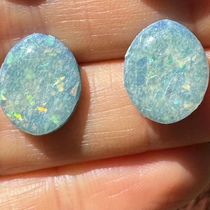 Puede incluir: Dos cabujones ovalados de ópalo azul iridiscente con fondo azul claro y motas blancas y doradas.