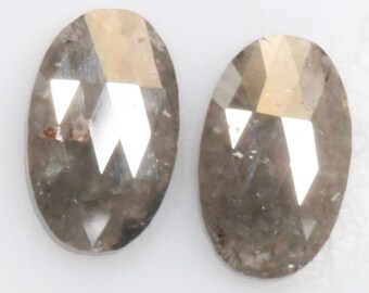 Pode incluir: Dois diamantes cinzentos de forma oval com corte facetado. Os diamantes têm um tom castanho claro e estão colocados sobre um fundo branco.
