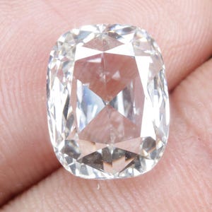 Puede incluir: Un diamante transparente de talla cojín con múltiples facetas, sostenido en la mano de una persona. El diamante refleja la luz, mostrando su brillo. La gema es de un color claro y brillante.