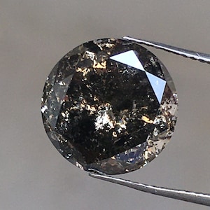 Może przedstawiać: Okrągły, ciemnoszary diament z czarnymi inkluzjami. Diament jest trzymany przez pęsetę.