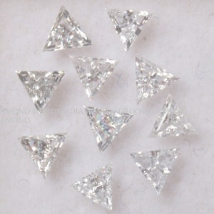 May include: A collection of twelve clear triangular diamonds, also known as trilliant cut diamonds, arranged in a circular pattern on a white surface.