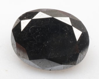 May include: A black, oval-cut gemstone with multiple facets. The polished surface reflects light, creating a sparkling effect. The gem is set against a white background. The stone is likely for jewellery.