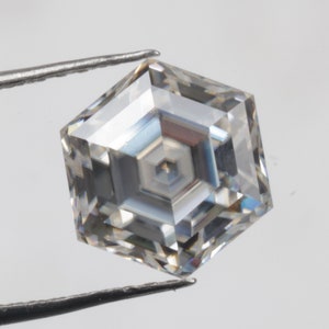 May include: A clear, faceted, hexagonal-shaped gemstone with a complex internal structure, held by tweezers against a white background.