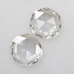 Puede incluir: Dos piedras preciosas redondas, transparentes y facetadas con un estilo de talla rosa, que presentan una base plana y una superficie superior abovedada y multifacetada que refleja la luz. Las piedras son incoloras y muestran facetas geométricas.