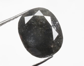May include: A dark grey oval-shaped gemstone with a white star-shaped pattern inside. The gemstone is held by a pair of tweezers.