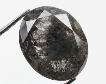 May include: A dark grey oval-shaped diamond with a faceted cut. The diamond is held by tweezers against a white background.