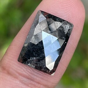 May include: A faceted, rectangular-cut gemstone with a dark gray color. The gem is held between two fingers, showcasing its reflective surfaces and intricate cuts. The background is blurred, with green foliage visible.