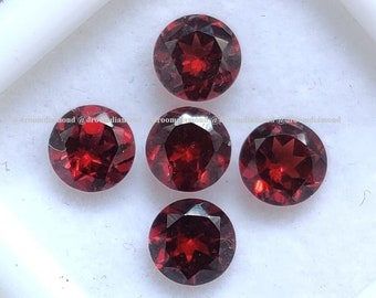 Puede incluir: Cinco piedras preciosas redondas de color rojo con un corte facetado. Las piedras preciosas están dispuestas en semicírculo sobre una superficie blanca.