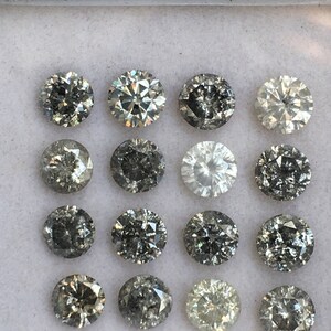 May include: A collection of 16 round, faceted, grey diamonds in a white box. The diamonds are arranged in four rows of four.