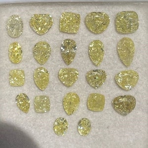 May include: A collection of 18 loose yellow diamonds of various shapes and sizes, including round, pear, heart, and square, displayed on a white surface.