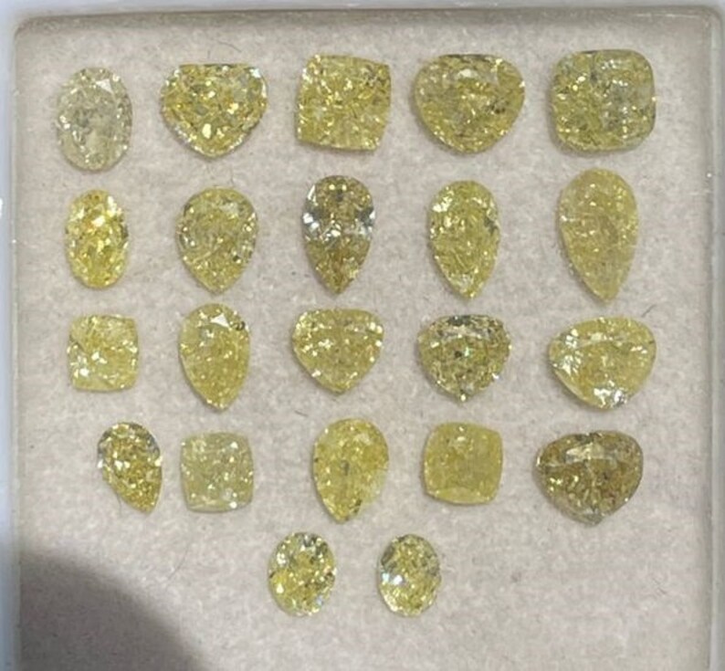 May include: A collection of 18 loose yellow diamonds of various shapes and sizes, including round, pear, heart, and square, displayed on a white surface.