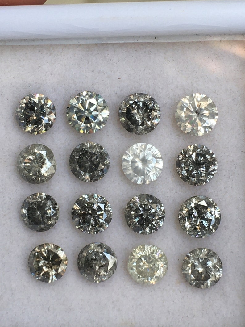 May include: A collection of 16 round, faceted, gray diamonds in a white box. The diamonds are arranged in four rows of four.