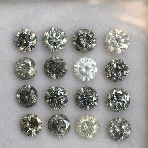 May include: A collection of 15 round, faceted, black diamonds in a white box. The diamonds are arranged in three rows of five.