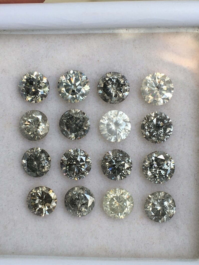 May include: A collection of 15 round, faceted, black diamonds in a white box. The diamonds are arranged in three rows of five.