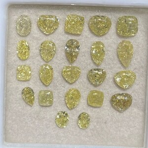 May include: A collection of 20 loose yellow diamonds of various shapes and sizes, including round, pear, square, and heart shapes. The diamonds are displayed in a white tray with a light brown background.