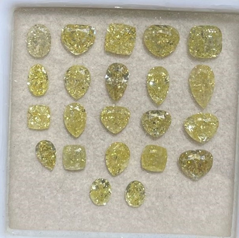 May include: A collection of 20 loose yellow diamonds of various shapes and sizes, including round, pear, square, and heart shapes. The diamonds are displayed in a white tray with a light brown background.