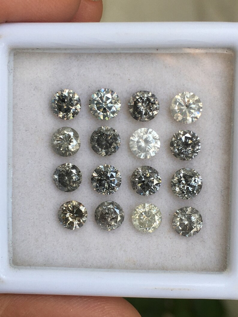 May include: A collection of 15 round, faceted, gray diamonds in a white box. The diamonds are arranged in three rows of five.