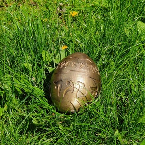 May include: A gold-colored egg with intricate designs and lettering on the surface, resting in a bed of green grass.