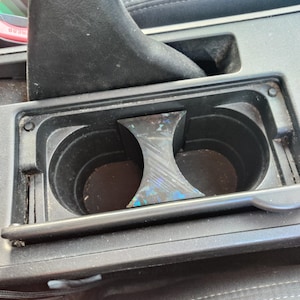 May include: A black car console cup holder with a central divider. The divider has a multi-coloured, iridescent finish. The cup holders are circular and empty. The console is made of black plastic.