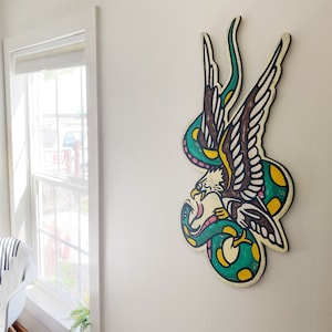 May include: A green and yellow snake wrapped around a brown and white eagle with pink accents. The eagle and snake are painted on a wooden surface.