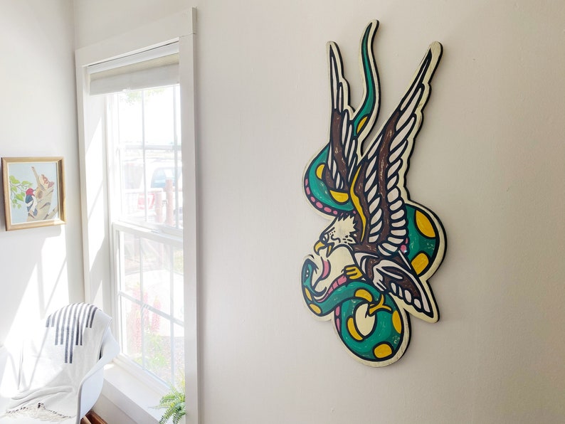 May include: A green and yellow snake wrapped around a brown and white eagle with pink accents. The eagle and snake are painted on a wooden surface.