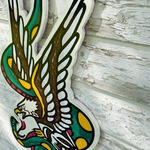May include: A green and yellow snake wrapped around a brown and white eagle with a pink beak. The eagle's wings are spread out and the snake's tail is curled around the eagle's leg. The image is painted on a wooden surface.