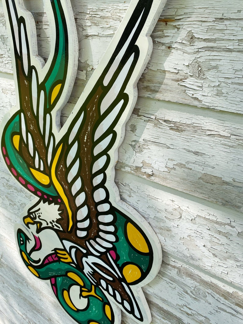 May include: A green and yellow snake wrapped around a brown and white eagle with a pink beak. The eagle's wings are spread out and the snake's tail is curled around the eagle's leg. The image is painted on a wooden surface.