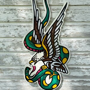 May include: A green and yellow snake wrapped around a brown and white eagle with pink accents. The eagle is attacking the snake. The image is a traditional tattoo design.