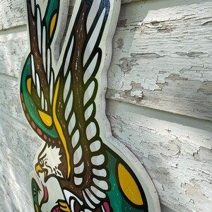 May include: A wooden wall hanging featuring a traditional American eagle tattoo design. The eagle is depicted with outstretched wings and a snake in its talons. The design is painted in green, yellow, brown, and white.