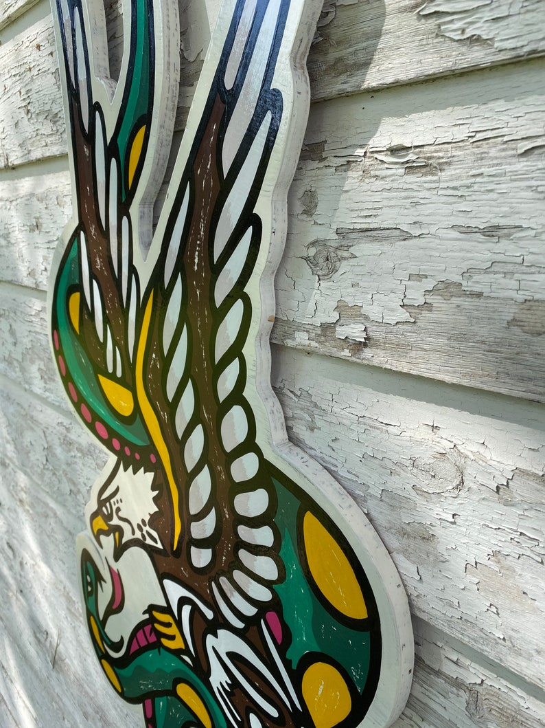 May include: A wooden wall hanging featuring a traditional American eagle tattoo design. The eagle is depicted with outstretched wings and a snake in its talons. The design is painted in green, yellow, brown, and white.