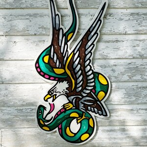 May include: A green snake wrapped around a brown eagle with yellow and pink accents. The eagle is flying with its wings spread. The image is painted on a wooden surface.
