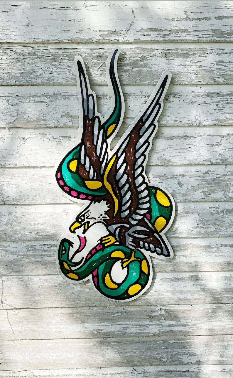 May include: A green snake wrapped around a brown eagle with yellow and pink accents. The eagle is flying with its wings spread. The image is painted on a wooden surface.