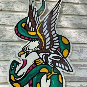 May include: A green snake with yellow spots wraps around a brown eagle with white feathers. The eagle's beak is open and it has a pink tongue. The image is painted on a white wooden surface.
