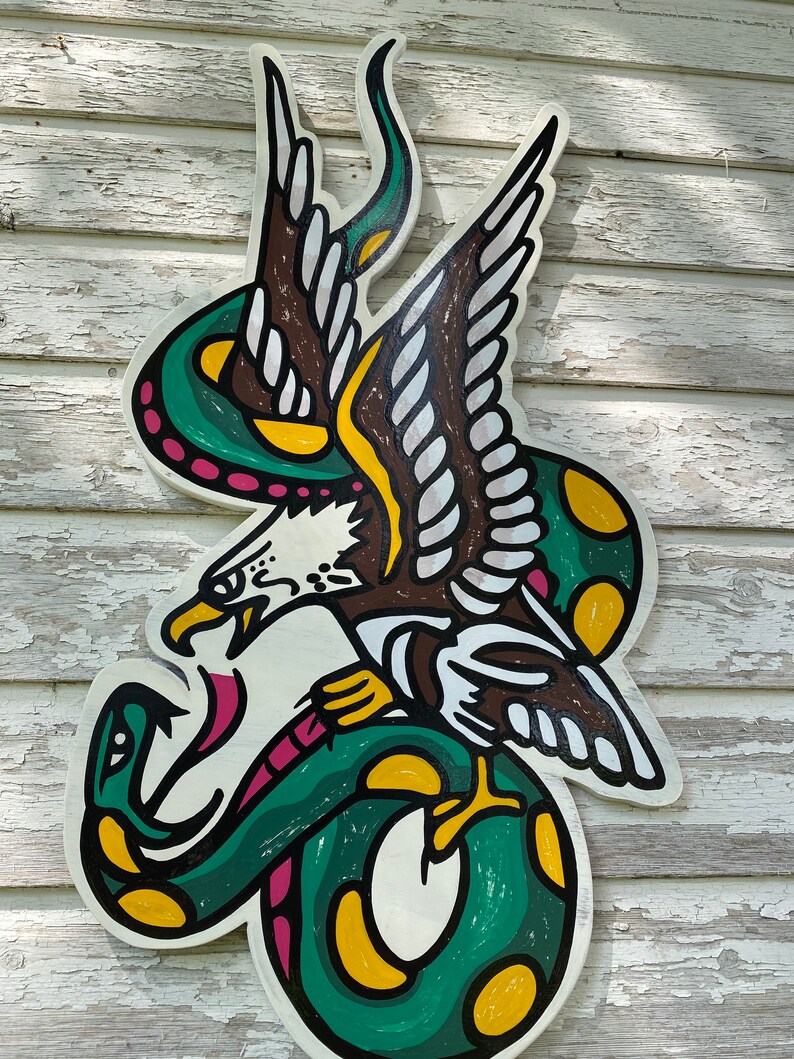 May include: A green snake with yellow spots wraps around a brown eagle with white feathers. The eagle's beak is open and it has a pink tongue. The image is painted on a white wooden surface.