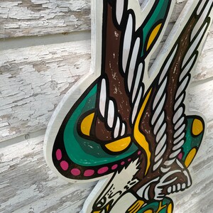 May include: A green, yellow, and pink eagle with wings outstretched, holding a green snake in its talons. The eagle is depicted in a traditional American tattoo style.