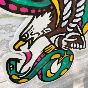 May include: A wooden cutout of an eagle with a snake in its talons. The eagle is painted white with black eyes and a pink beak. The snake is green with yellow spots and a pink tongue. The eagle's wings are green and yellow with pink dots.