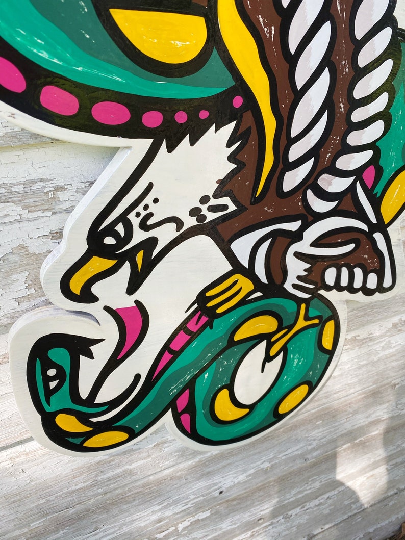 May include: A wooden cutout of an eagle with a snake in its talons. The eagle is painted white with black eyes and a pink beak. The snake is green with yellow spots and a pink tongue. The eagle's wings are green and yellow with pink dots.